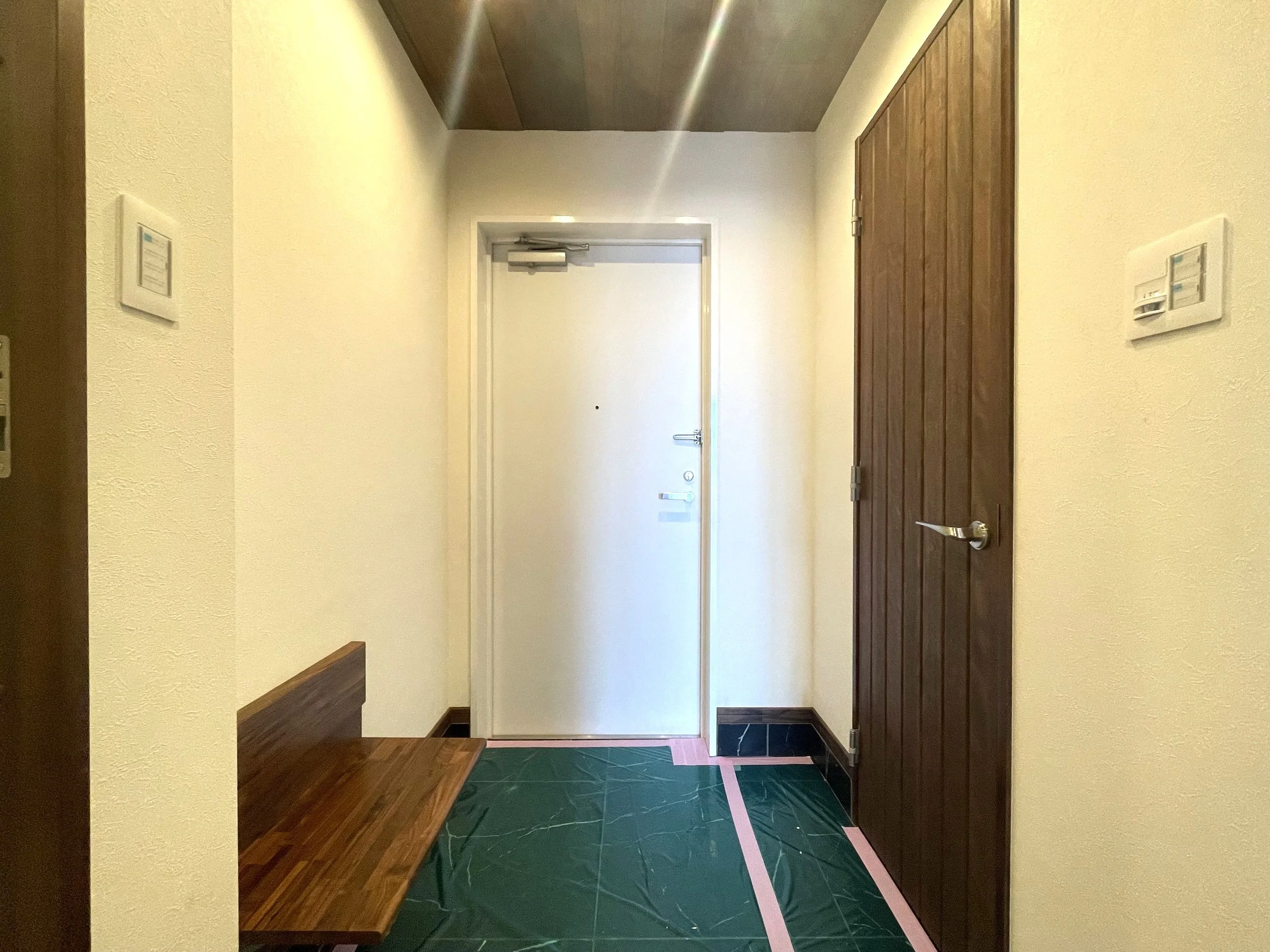 玄関のドアと壁、木製の扉、コンクリートの床の上に緑色の養生シートとピンク色の養生テープが貼られている室内の風景