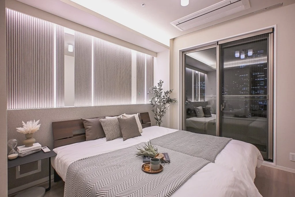 モダンな寝室の内装で、ダブルベッド、クッション、植物、サイドテーブル、夜景が見えるガラスドアがあります。