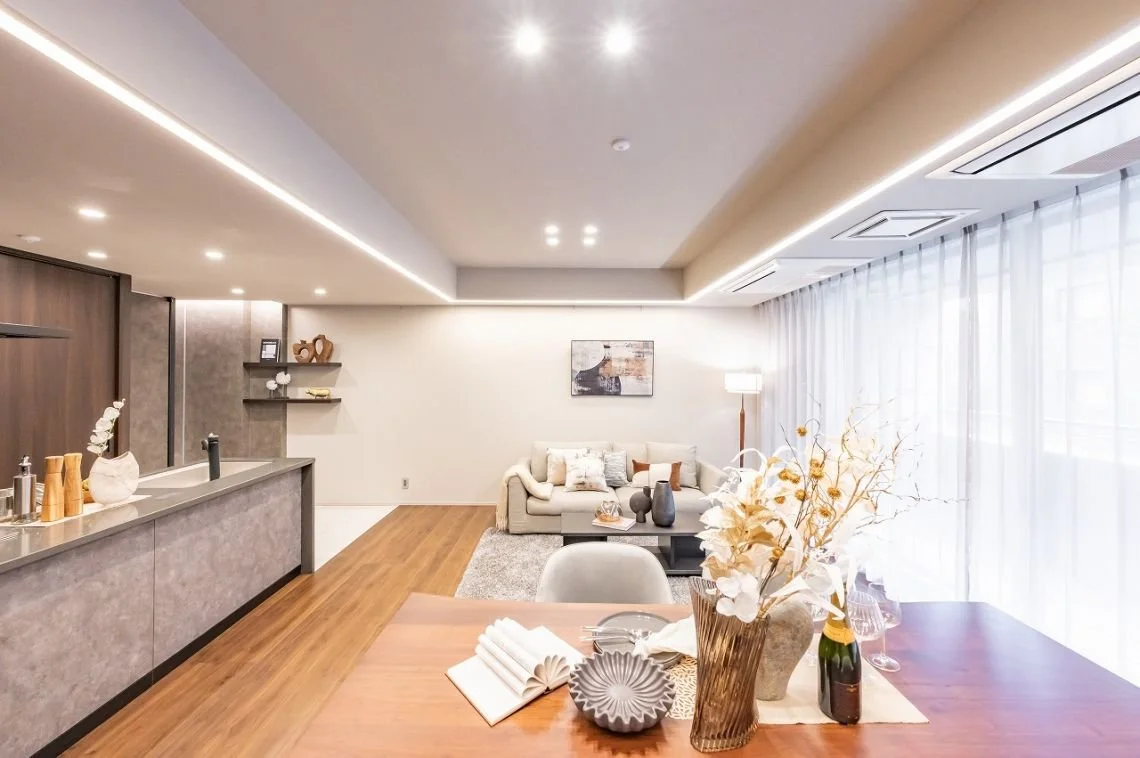 明るいリビングルームとダイニングエリア。リビングには白いソファとカーペット、壁に絵画。ダイニングには花瓶とテーブルインテリア。窓からの自然光。