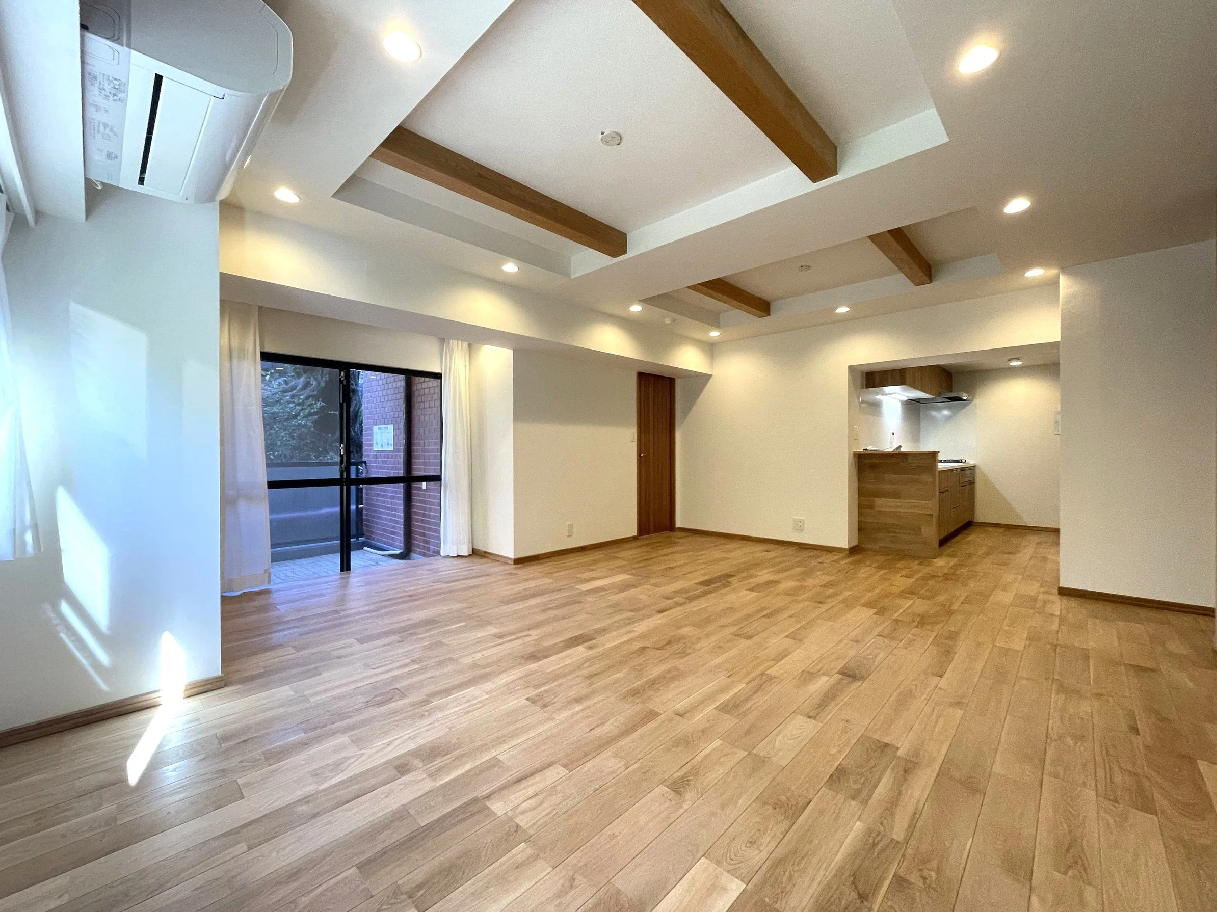 木の床と天井に木の梁がある広いリビングルーム。窓側には白いカーテン、スライドドアを通じてバルコニーが見える。キッチンが奥にあり、壁と天井は白色。照明も点灯している。