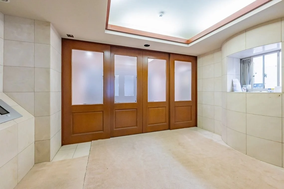 木製のドアが3つ並んでいる玄関ホール。左側は壁に埋め込まれたインターホンらしきものがある。右側には窓や棚があり、窓にはカーテンといくつかの小物が置かれている。