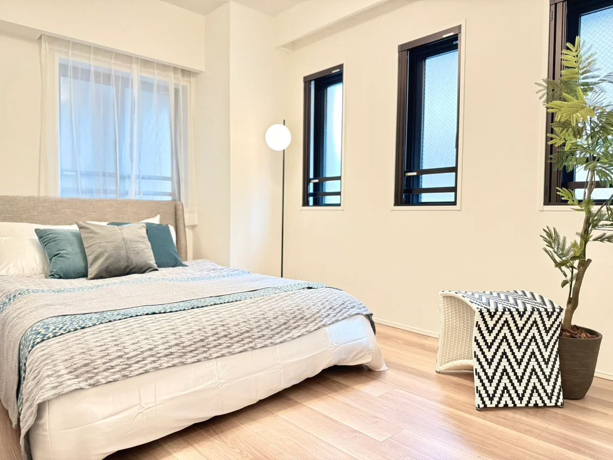 シンプルなベッドルームで、ベッドにクッションが置かれている。横には観葉植物と模様のあるスツールがある。窓にはカーテンとブラインドがあり、間接照明のスタンドも置かれている。