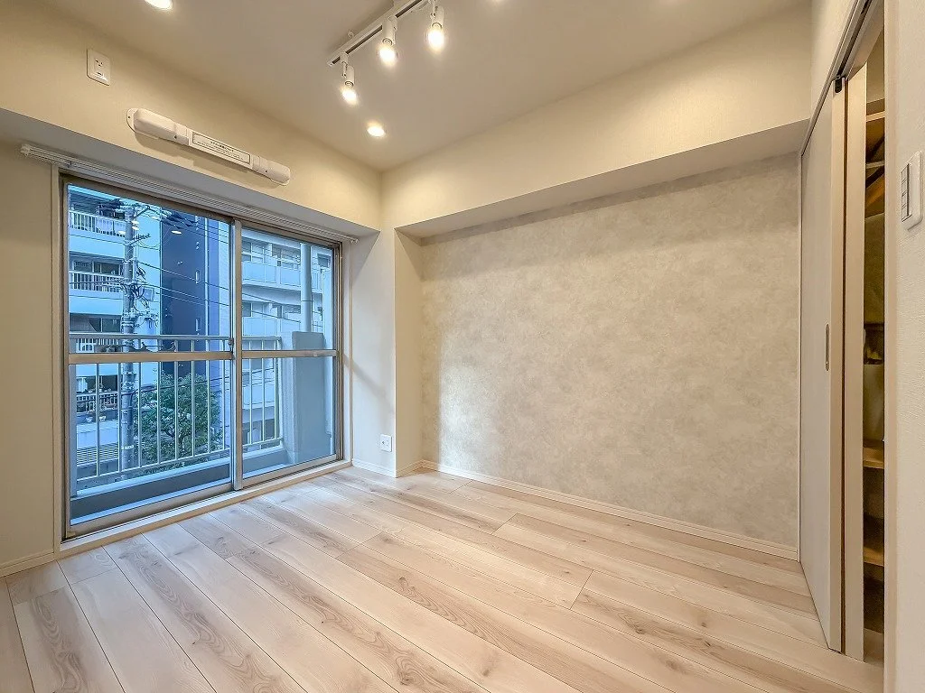 明るい空間の部屋で、大きな窓から都市の建物が見える。天井に照明とエアコンが設置されている。床は木製。壁は淡い色合い。