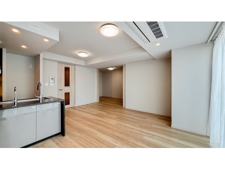 空のリビングルームとキッチンエリアの写真。木製の床と白い壁、窓からの自然光が入っている。」}}