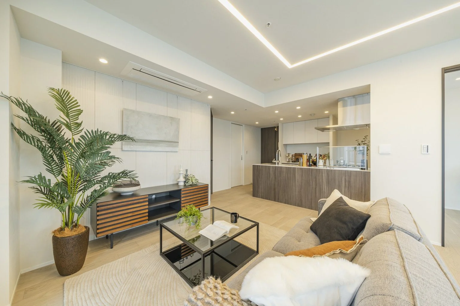モダンなリビングルームとキッチンのインテリア。大きな観葉植物、ソファ、ガラスのコーヒーテーブル、サイドボードが配置されている。