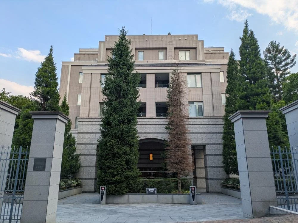都市のビルの正面、入口付近に大きな木と縁石があり、門柱に看板やセキュリティボックスが設置されている。