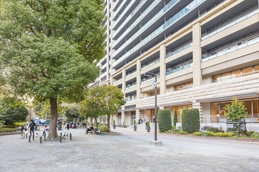 高層マンションの前の公園で、木やベンチ、時計台と人々がいる景色