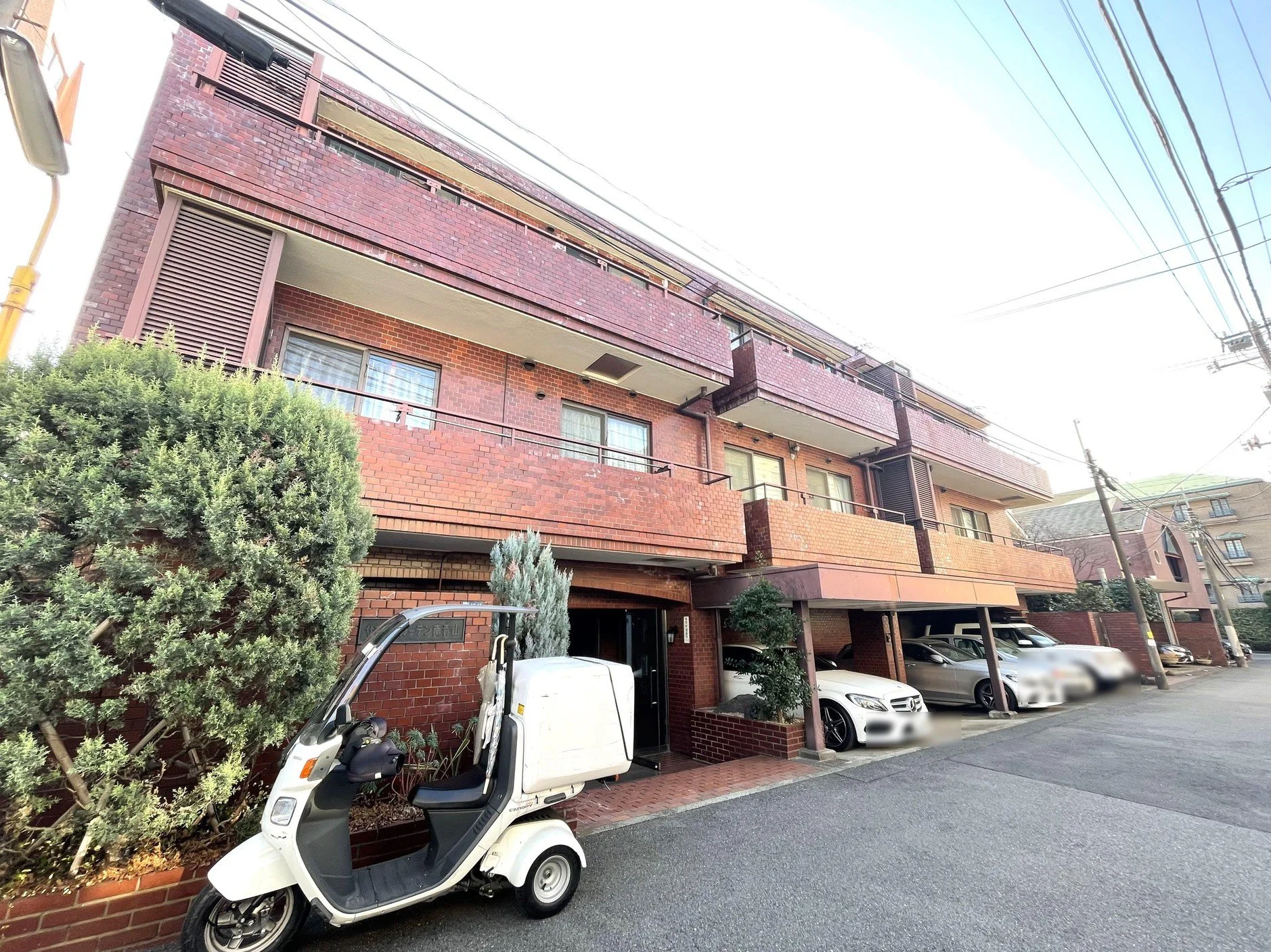 赤レンガのアパートメントまたはマンションの建物。駐車場に車とバイクが停車している。前景に緑の木と白いバイクが見える。電線が空に張り巡らされている。