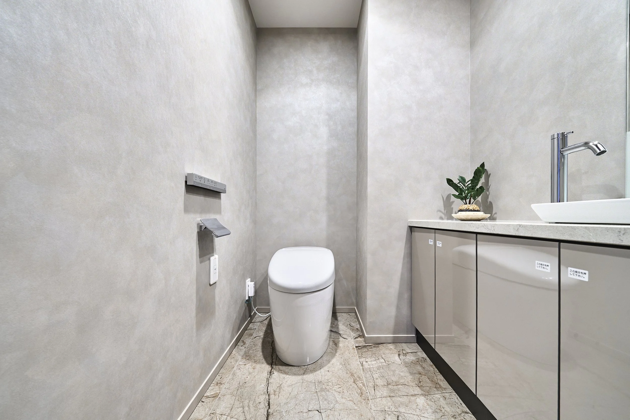 シンプルでモダンなトイレルーム。白い便器と洗面台、緑の観葉植物が置かれている。清潔感のある内装で、壁紙は淡い色調。