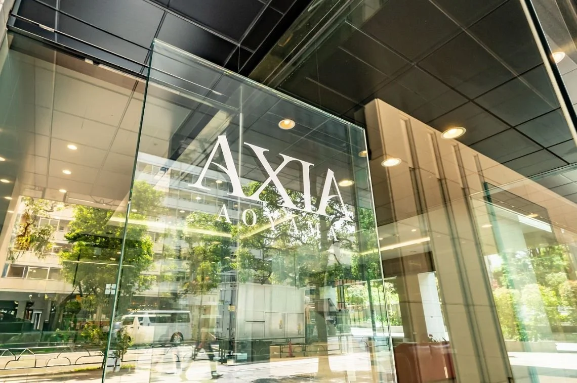 ガラスの壁に書かれた『AXXA MARINA』という文字。外の街並みと木々が反射して見える現代的な建物の入り口部分の写真。」}always consider accessibility by providing clear, concise, and descriptive alt text that captures the essential visual information of the image. If the image contains text, include it in 
