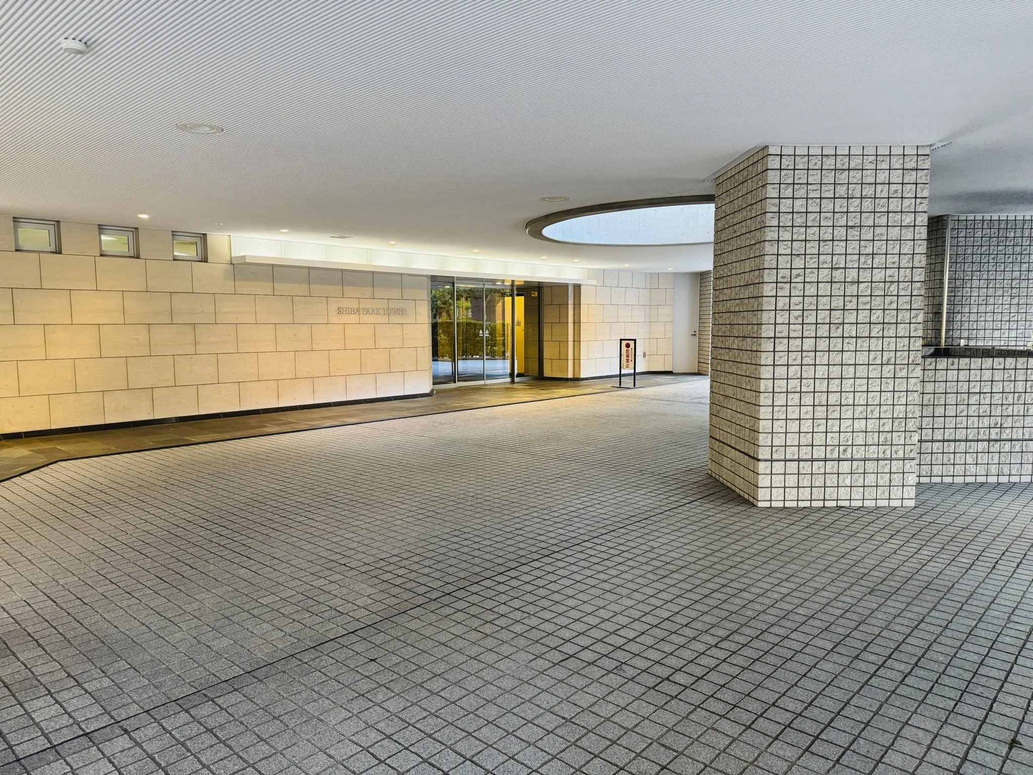建物の正面玄関前の明るい屋外エントランス。壁は白いレンガ調、地面は格子状のタイル。扉の上に「SHIBATA PARK TOWER」と書かれている。
