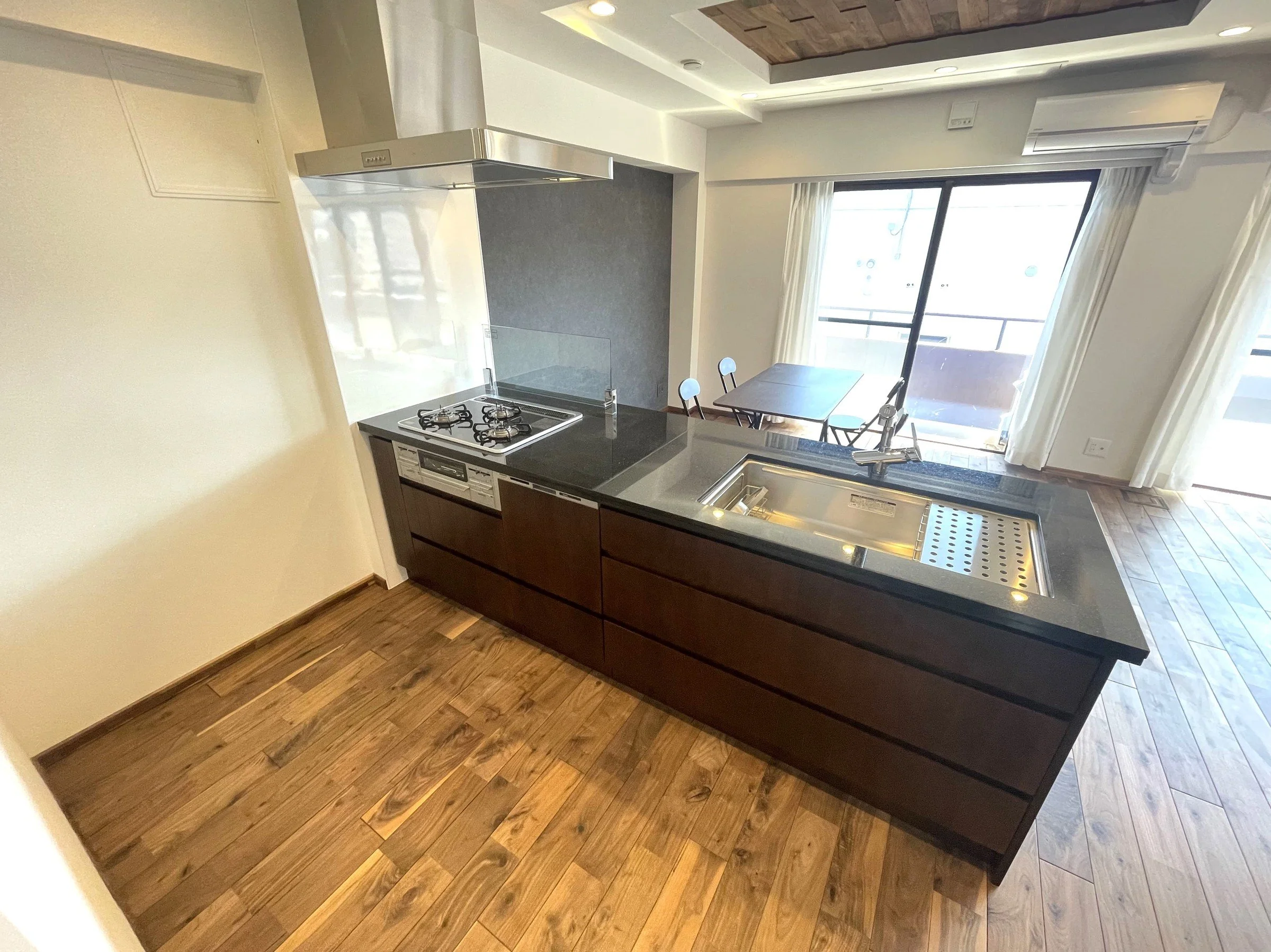 シンクとガスコンロが付いたキッチンとダイニングのスペースが見える部屋の写真。大きな窓から自然光が入り、バルコニーが見える。