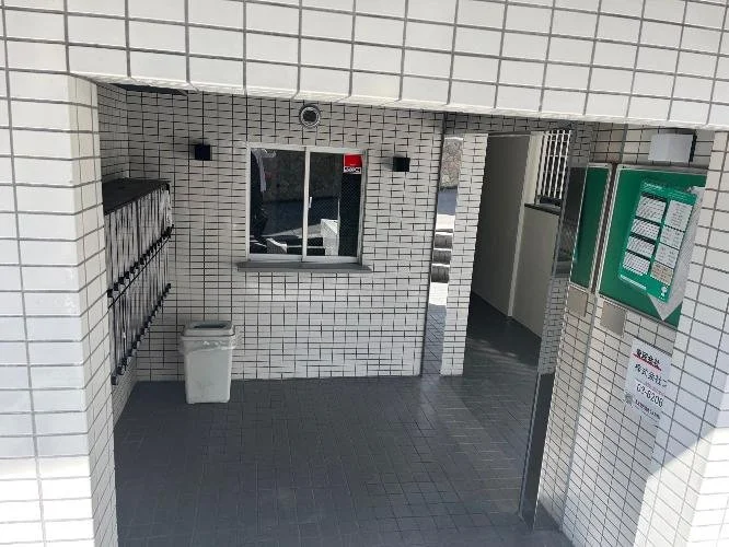 白いタイル張りの建物の入口付近。郵便受け、ゴミ箱、掲示板が設置されている。