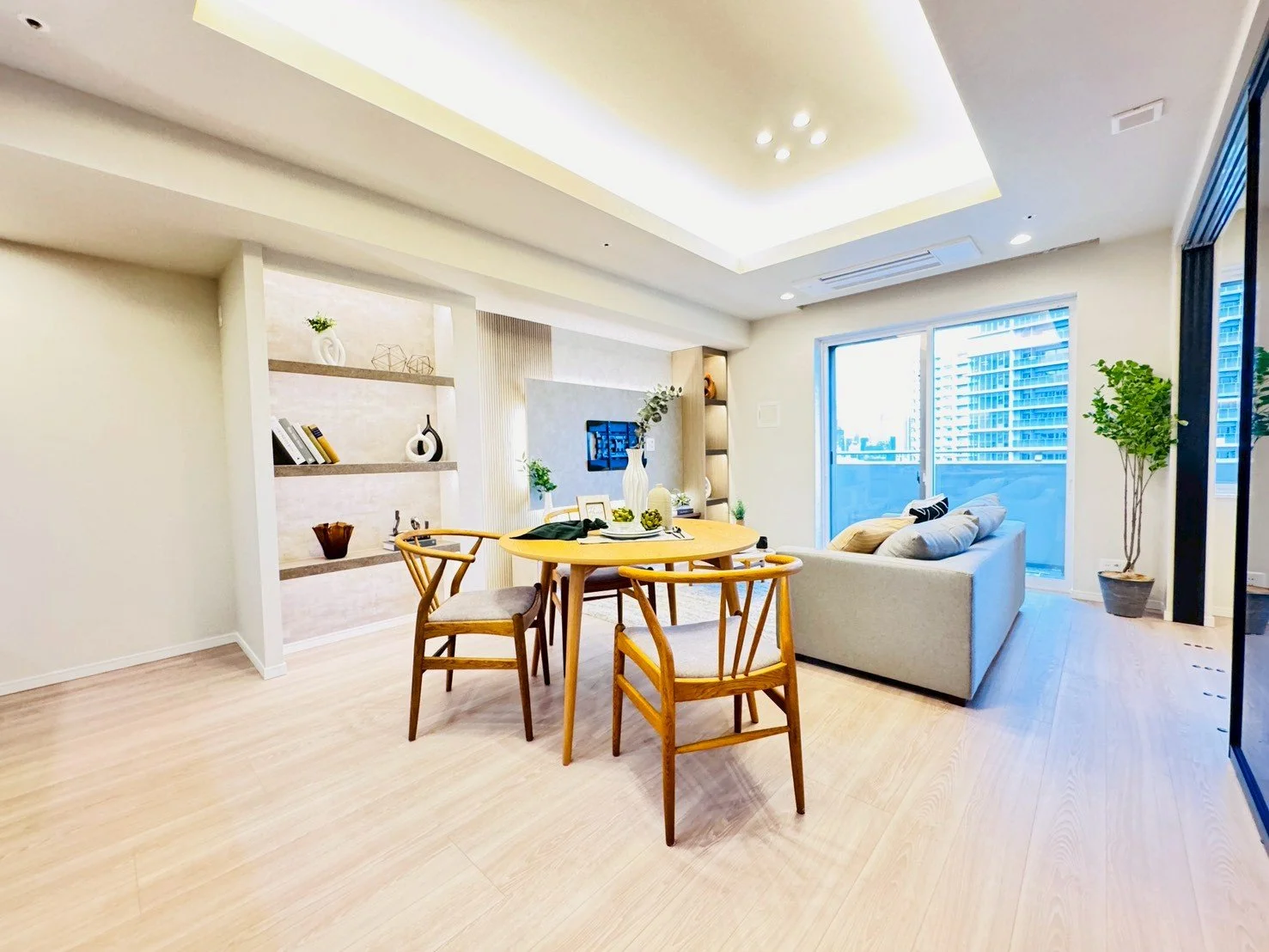 明るいリビングルーム。円形の木製テーブルと4脚の木製椅子、ソファと背もたれクッション、棚に飾りや本、観葉植物、大きな窓とバルコニーがあります。