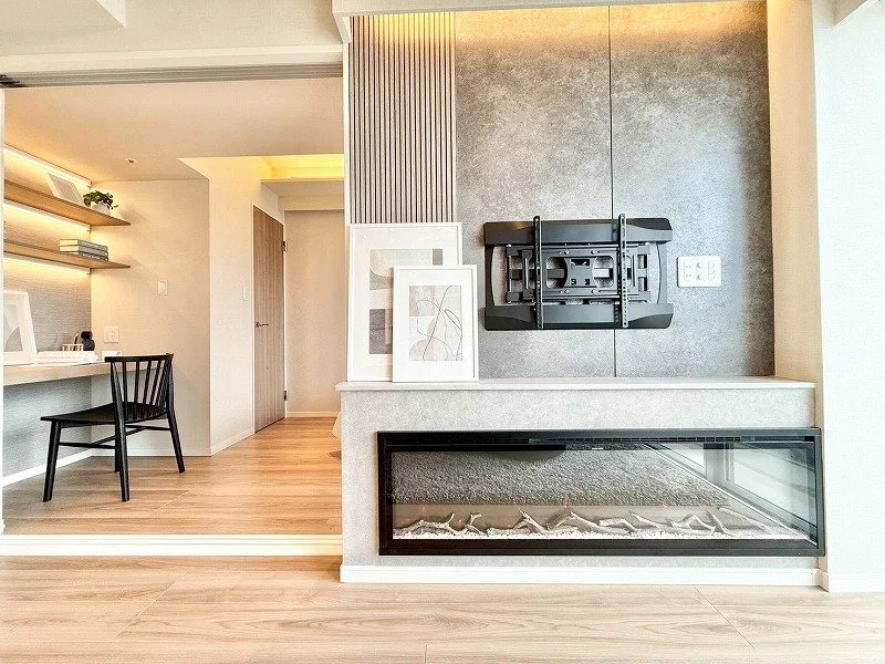 リビングルームの壁に掛けられたテレビのブラケットとテレビの壁掛け金具、ソファの下のガラス張りの暖炉、壁に飾られた抽象画、木製の床と壁にある本棚や椅子