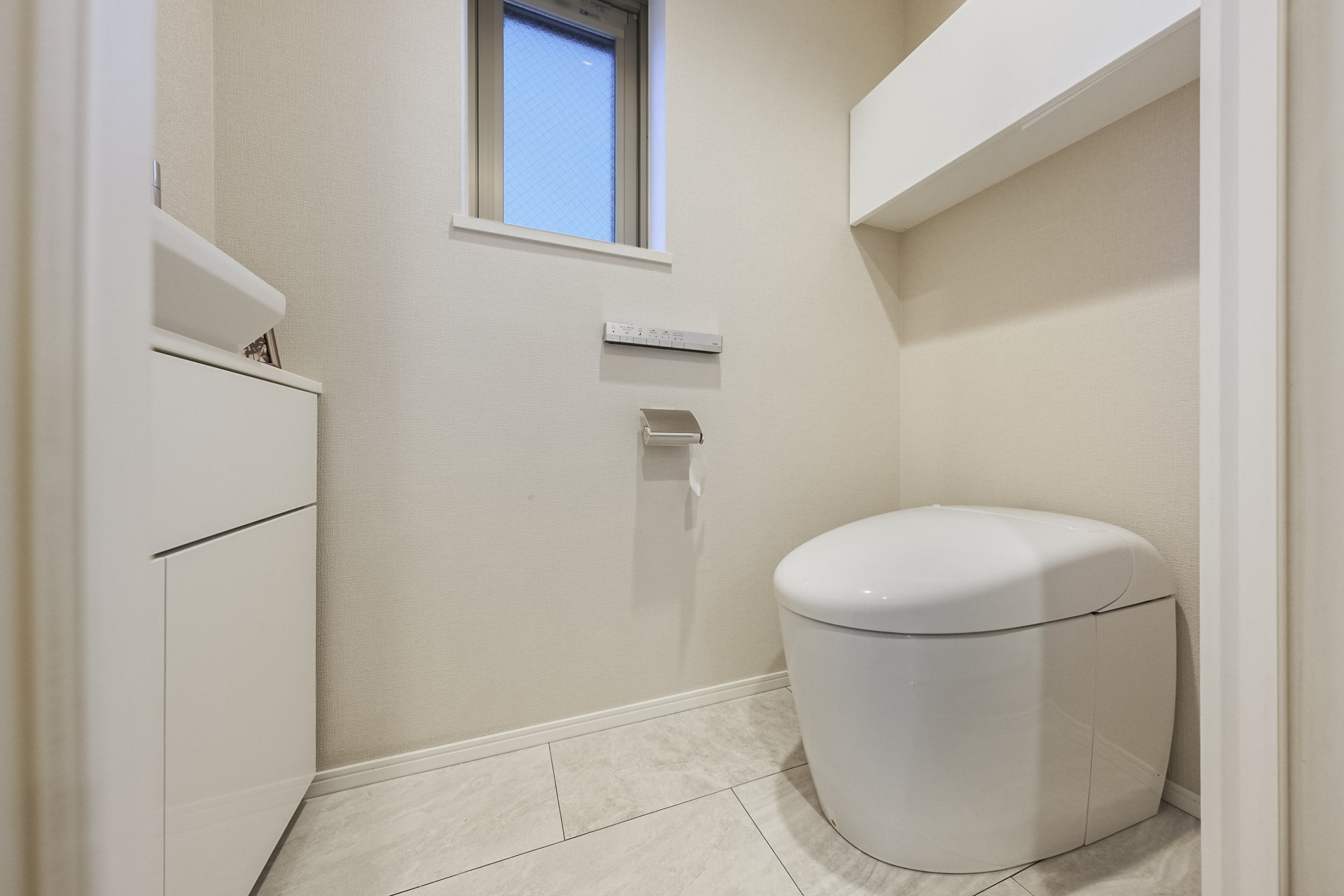 白い便座のトイレと小さな白い収納棚と窓のあるトイレルーム