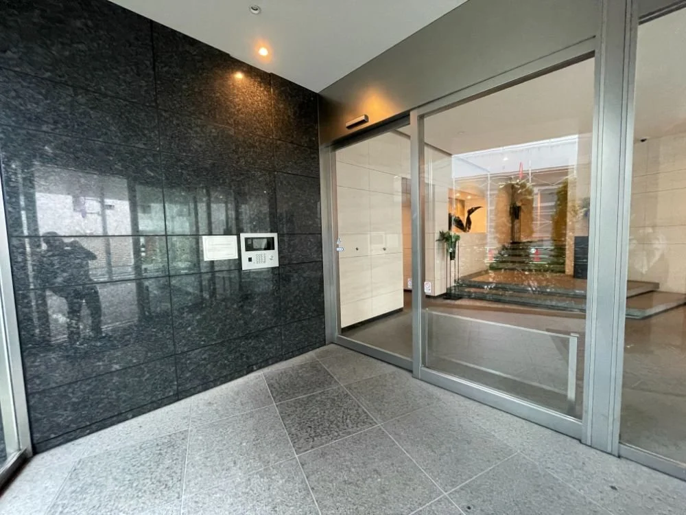建物のエントランスの内側のショット。黒い石の壁にインターホンとモニターが設置されている。ガラス扉を通じてエントランスホールが見える。