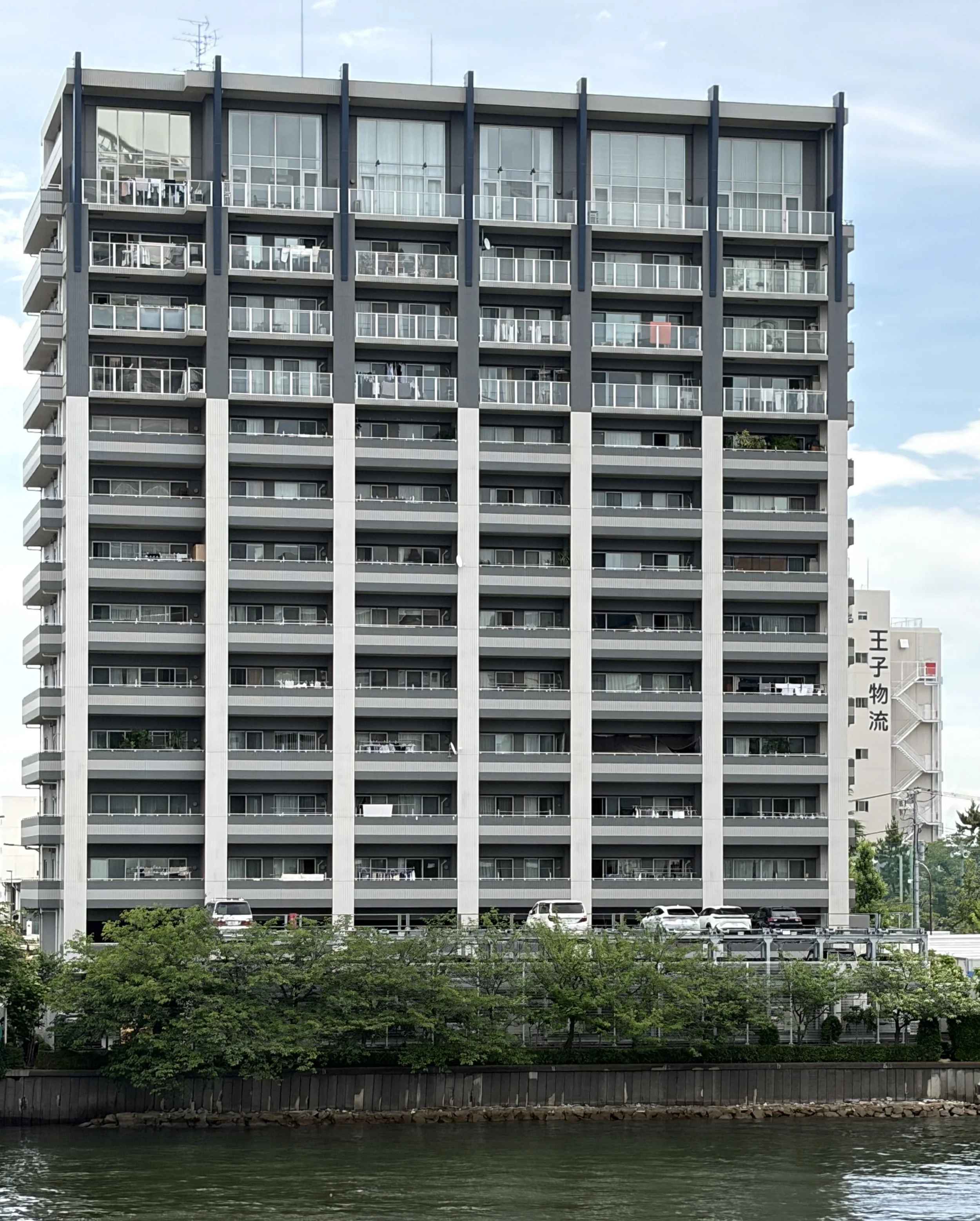 水辺に面した高層のコンドミニアムまたはオフィスビル。複数の階にわたってベランダや窓が並び、車が駐車されている。上部には空と一部雲、下部には木々と水が見える。