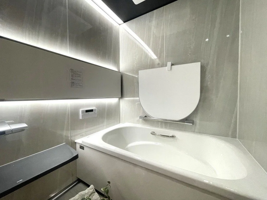 白いバスタブと回転して蓋を閉じるタイプの浴槽蓋が設置された浴室の写真。浴室壁は灰色のタイル張りで、照明が明るく照らしている。