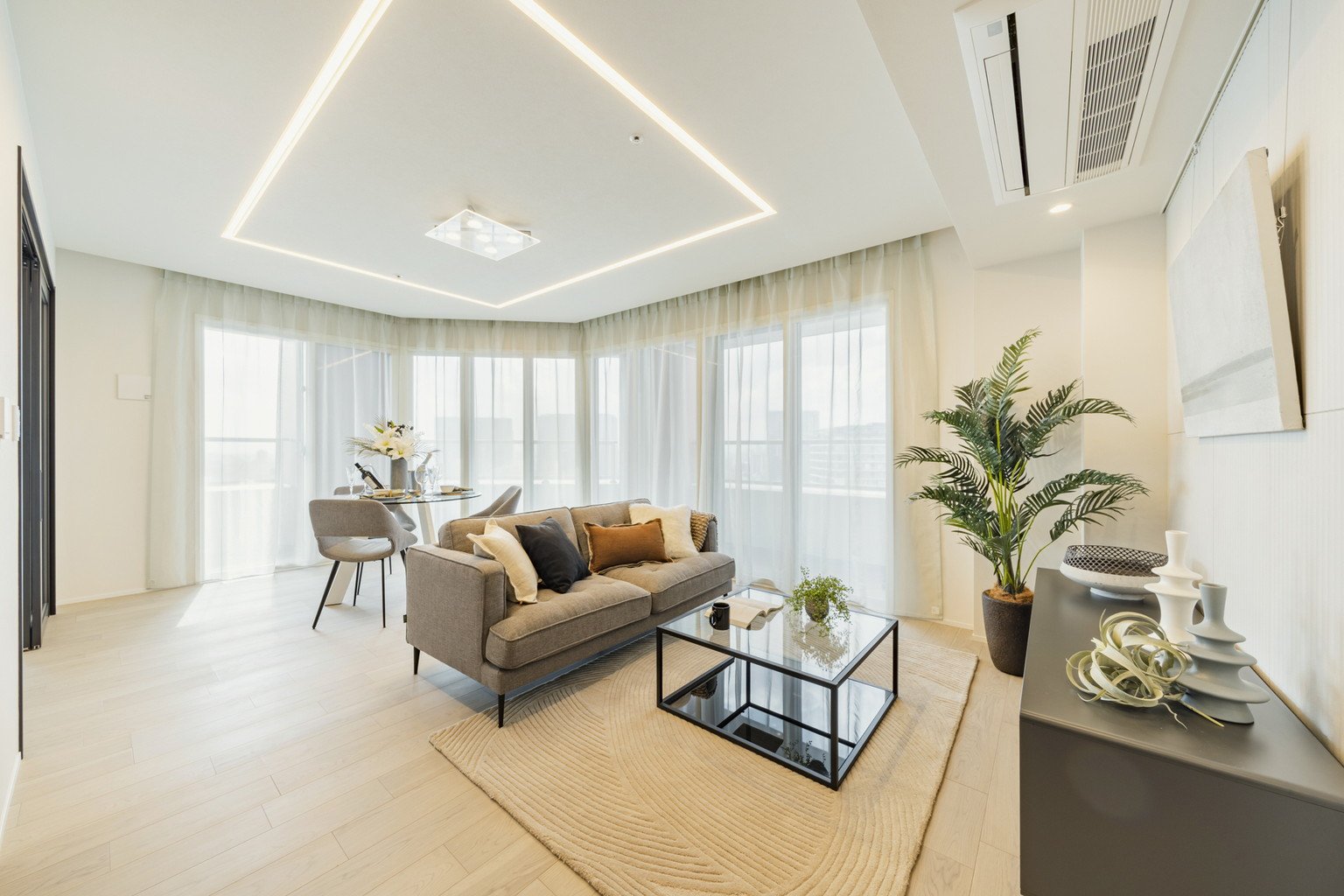 明るいリビングルームに灰色のソファとガラスのコーヒーテーブル、ダイニングセットと観葉植物が配置されている。窓側には白いカーテンがかかっている。