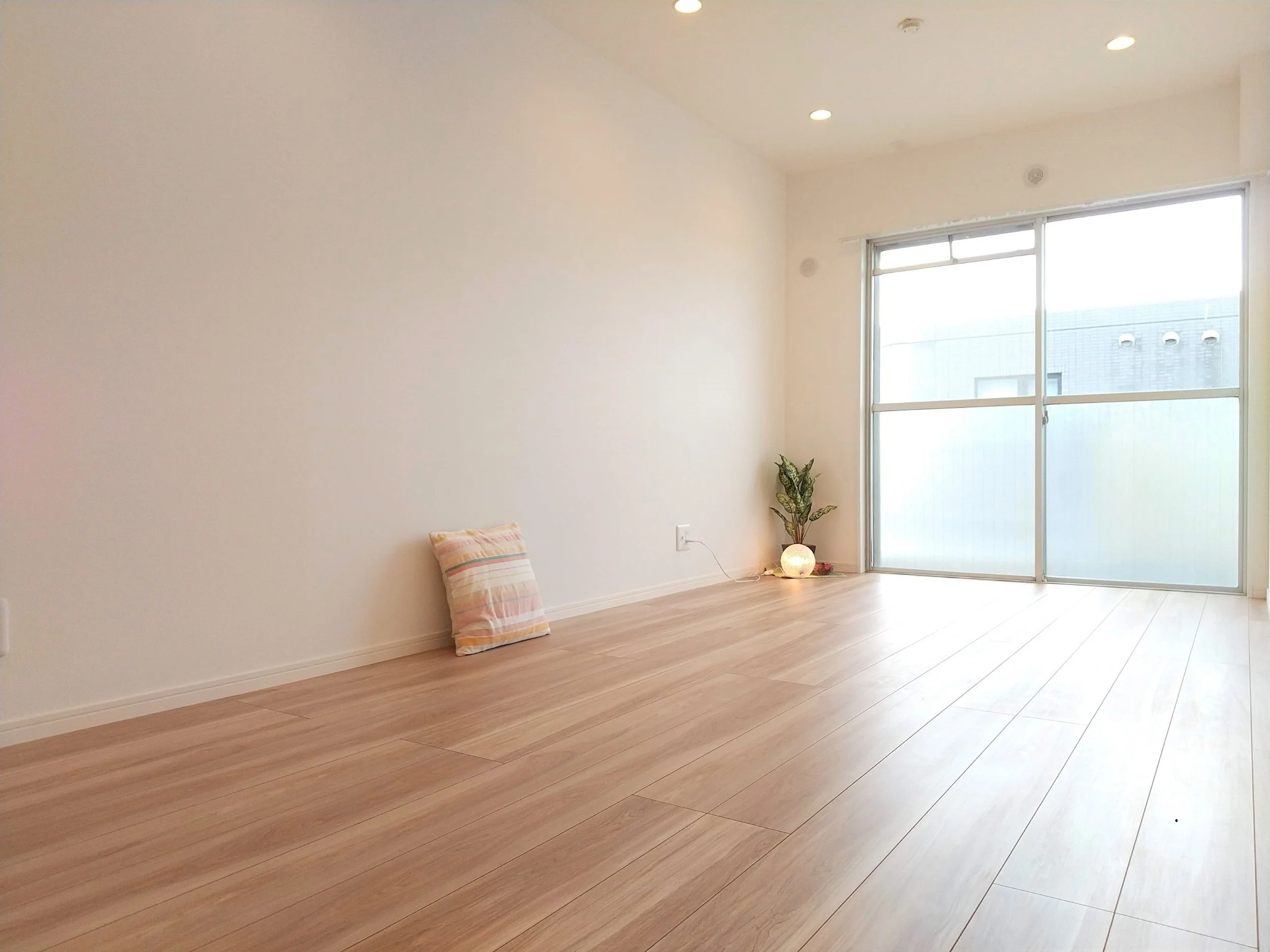 白い壁と木製の床があるシンプルな部屋。窓から自然光が差し込み、観葉植物とランプが置かれている。