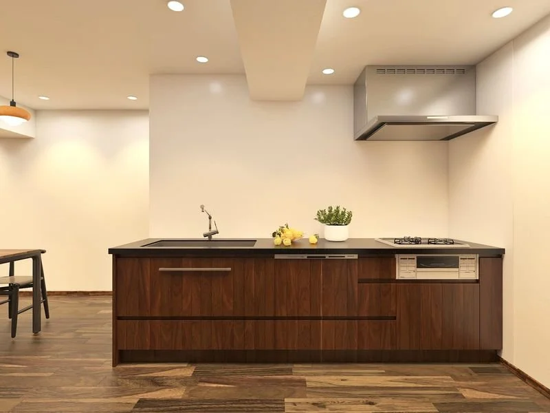 シンプルなモダンキッチンの画像。木製のキャビネットと黒いカウンター、シンク、ガスコンロと換気扇、観葉植物と黄色い花が飾られている。部屋は明るく広々としている。