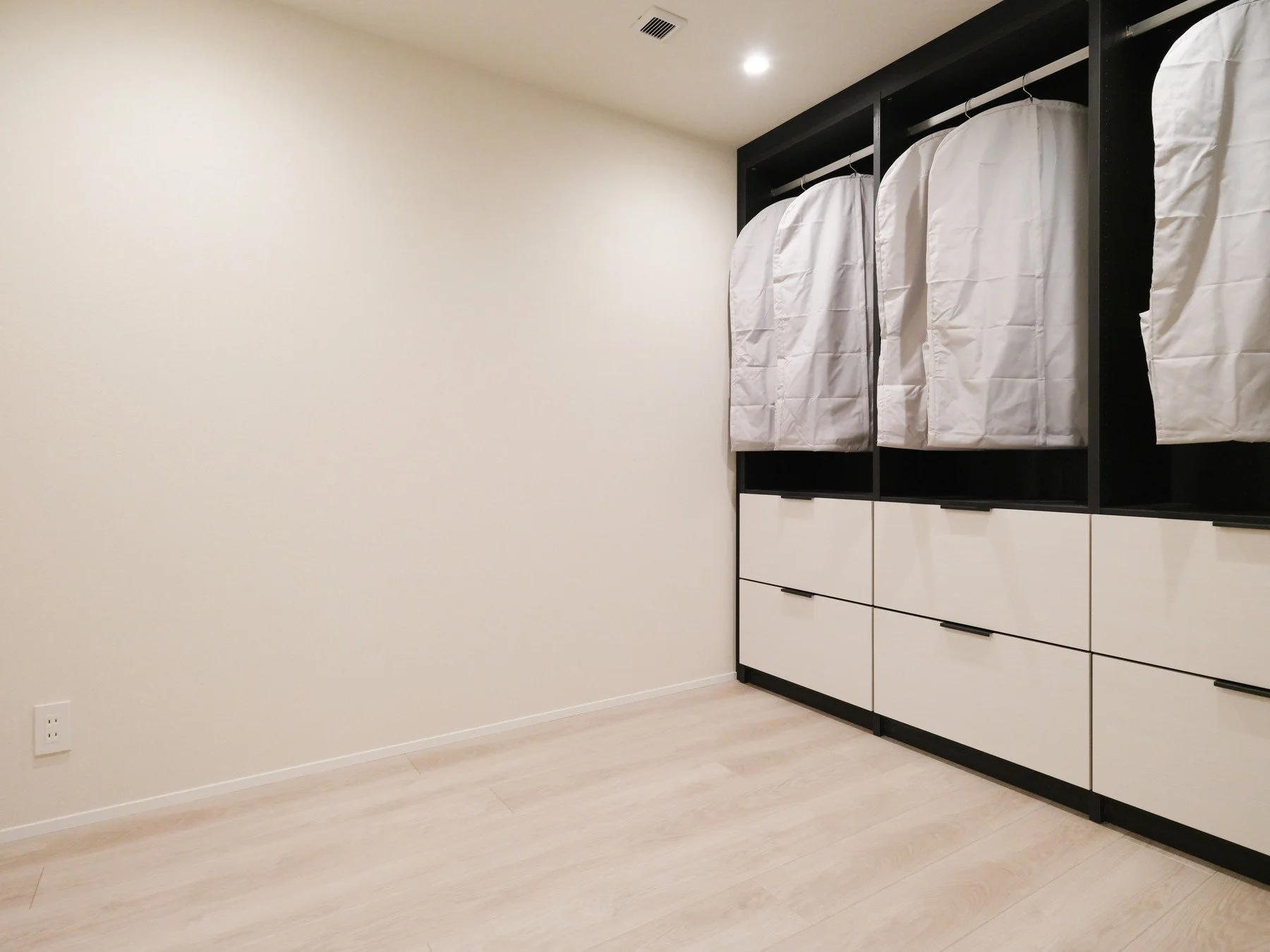 白い布巻きの衣装ケースが並ぶ棚が黒い壁に設置された部屋の一角、木製の床と空白の壁が見える