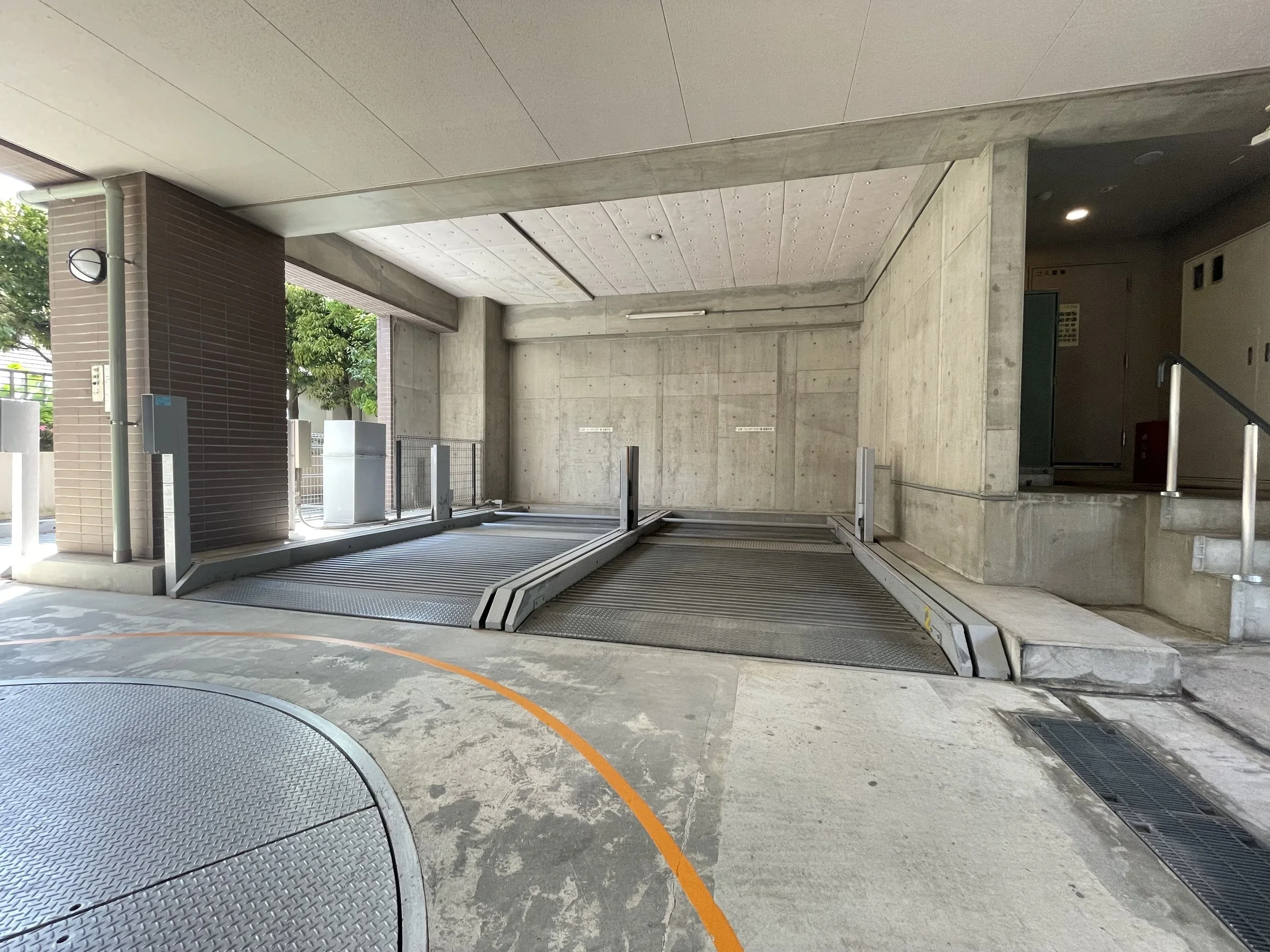 コンクリート造りの駐車場ゲート。自動スロープ式駐車場ゲートと昇降装置。周囲に金属製の柵と排水溝が見える