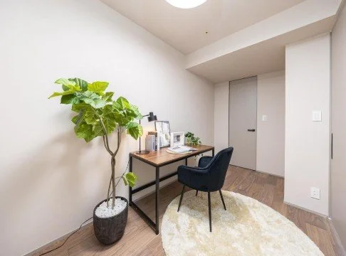 白い壁の部屋にデスクと椅子、観葉植物が置かれている。窓やドアも見えるシンプルな室内風景.