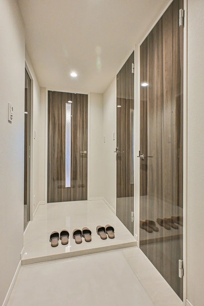 玄関のホールで、スリッパが三足置かれている。背面には木目調のドアがあり、その横にはガラス扉がある。