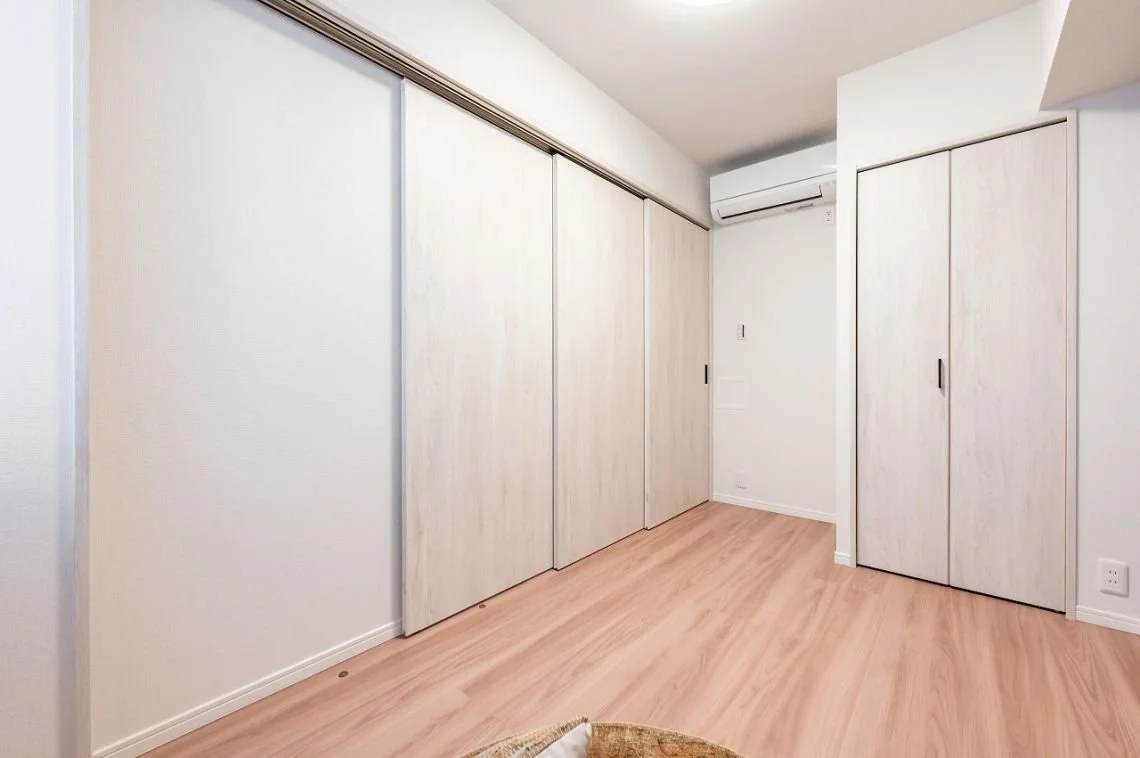 白い壁と木製の折戸式収納ドアのある部屋の一角。床は木のフローリングで、エアコンの室外機が設置されている。