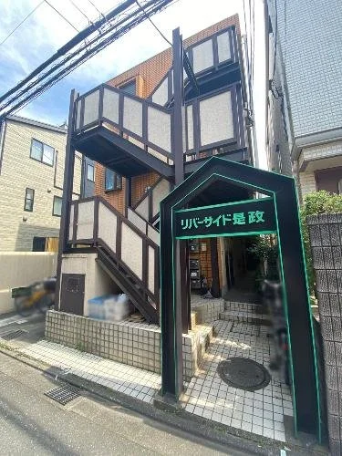 三階建ての建物の入り口に、緑色の照明付きの看板と階段があり、周囲には住宅や電線が見える都市の風景です。