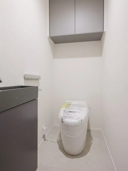 白い壁のトイレルーム、便器と上に収納棚があるシンプルな空間。