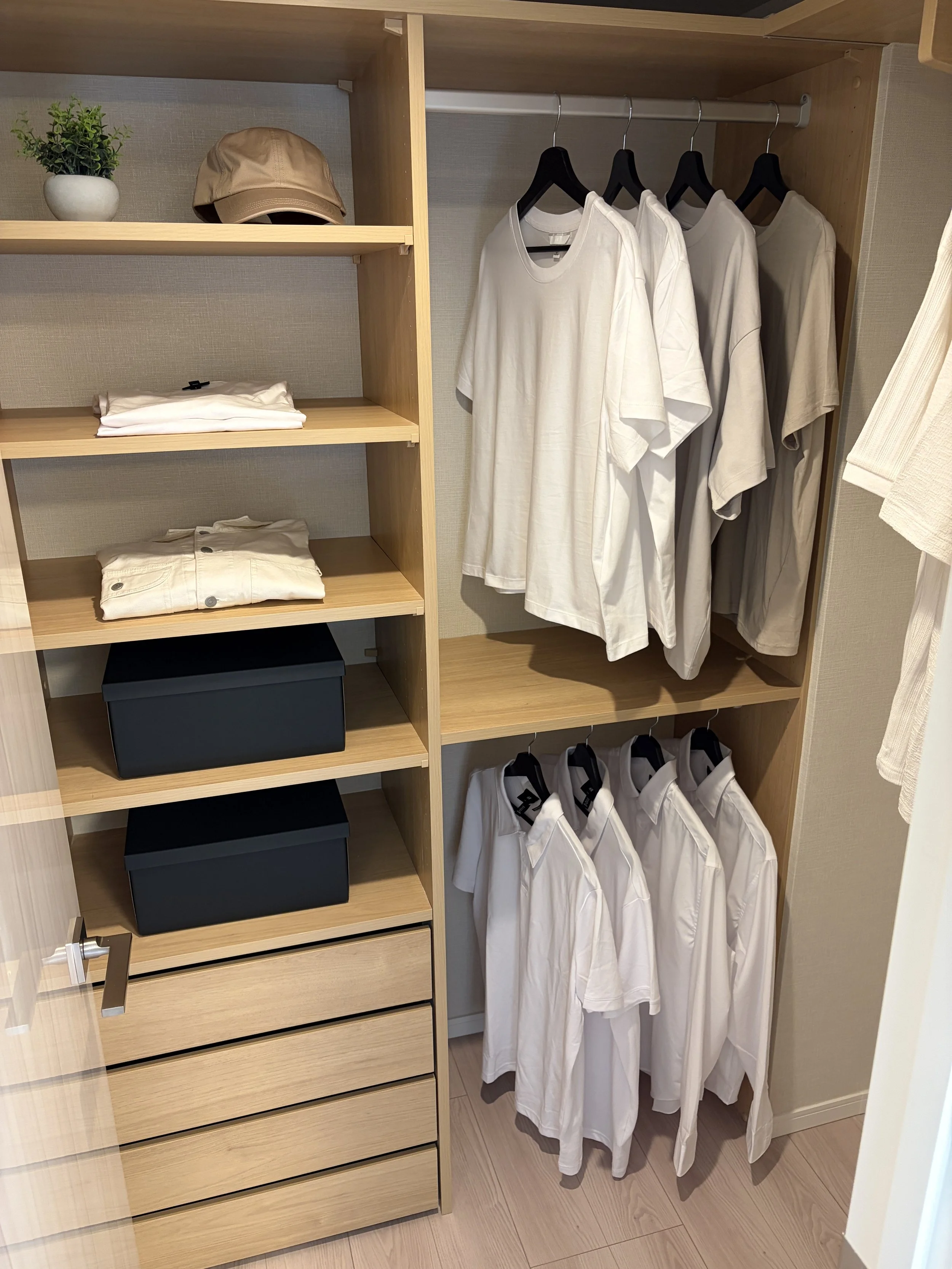 木製のクローゼットの中に白いTシャツやシャツがかかっている。左側には棚があり、上段には植物とベージュの帽子、下段には白いシャツと黒い箱が収納されている。