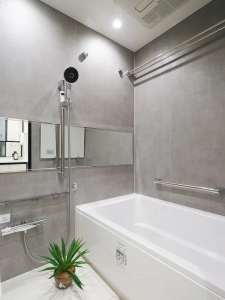 モダンなバスルームの浴室。壁はグレーのタイルで、洗い場にシャワーと大きな浴槽があり、鏡と緑の観葉植物も見える。