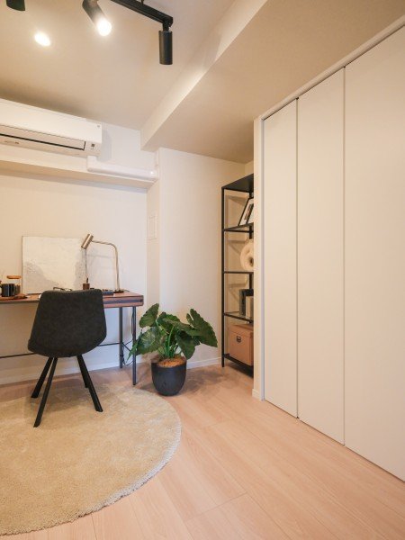 白い壁と木製の flooring の部屋。デスクと椅子、観葉植物、収納棚が見えるシンプルな家具の配置。
