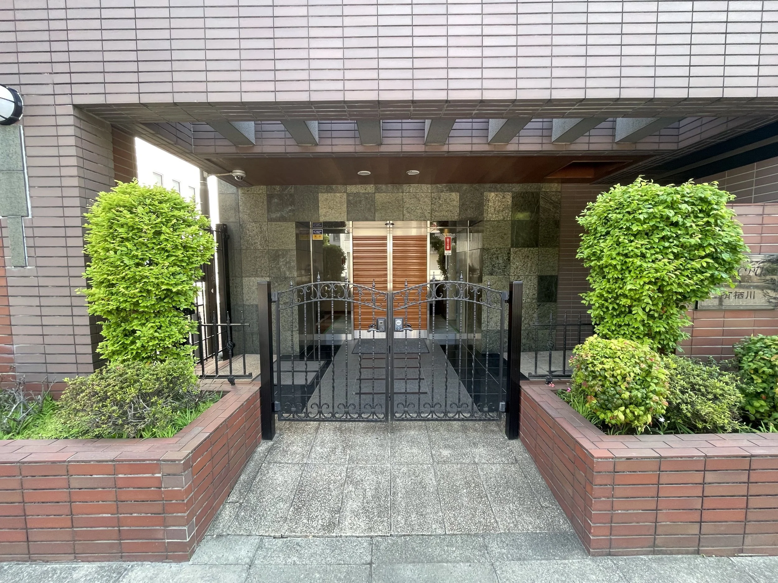 黒い鉄の門の前に緑の生垣と低い花壇があり、ビルの入口に向かっている。