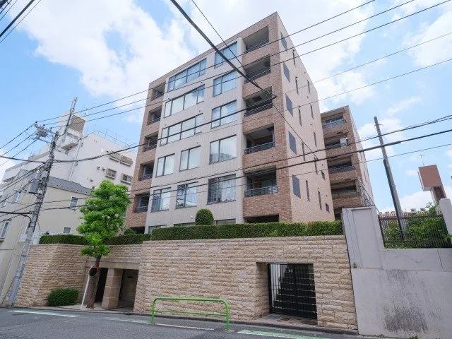六階建ての現代的なマンションの外観。レンガとコンクリートの建物で、複数のバルコニーと大きな窓が特徴。前には小さな木と緑の生垣、フェンスがあり、道路に面している。