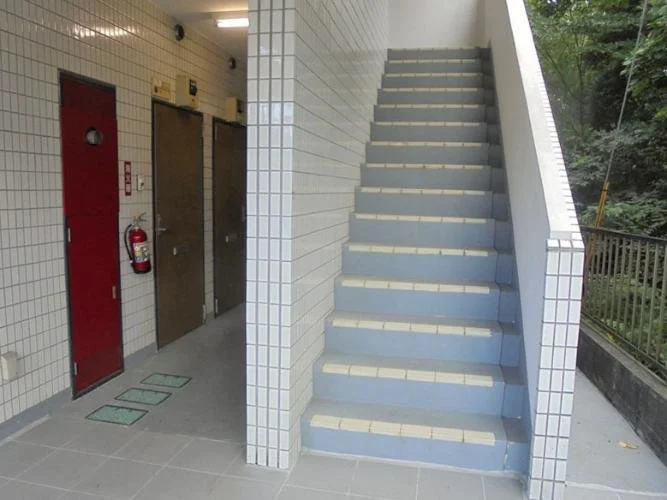 白いタイル張りの壁と階段がある建物内の廊下。階段は外に続いており、左側にエレベーターと火災警報器が設置されている。