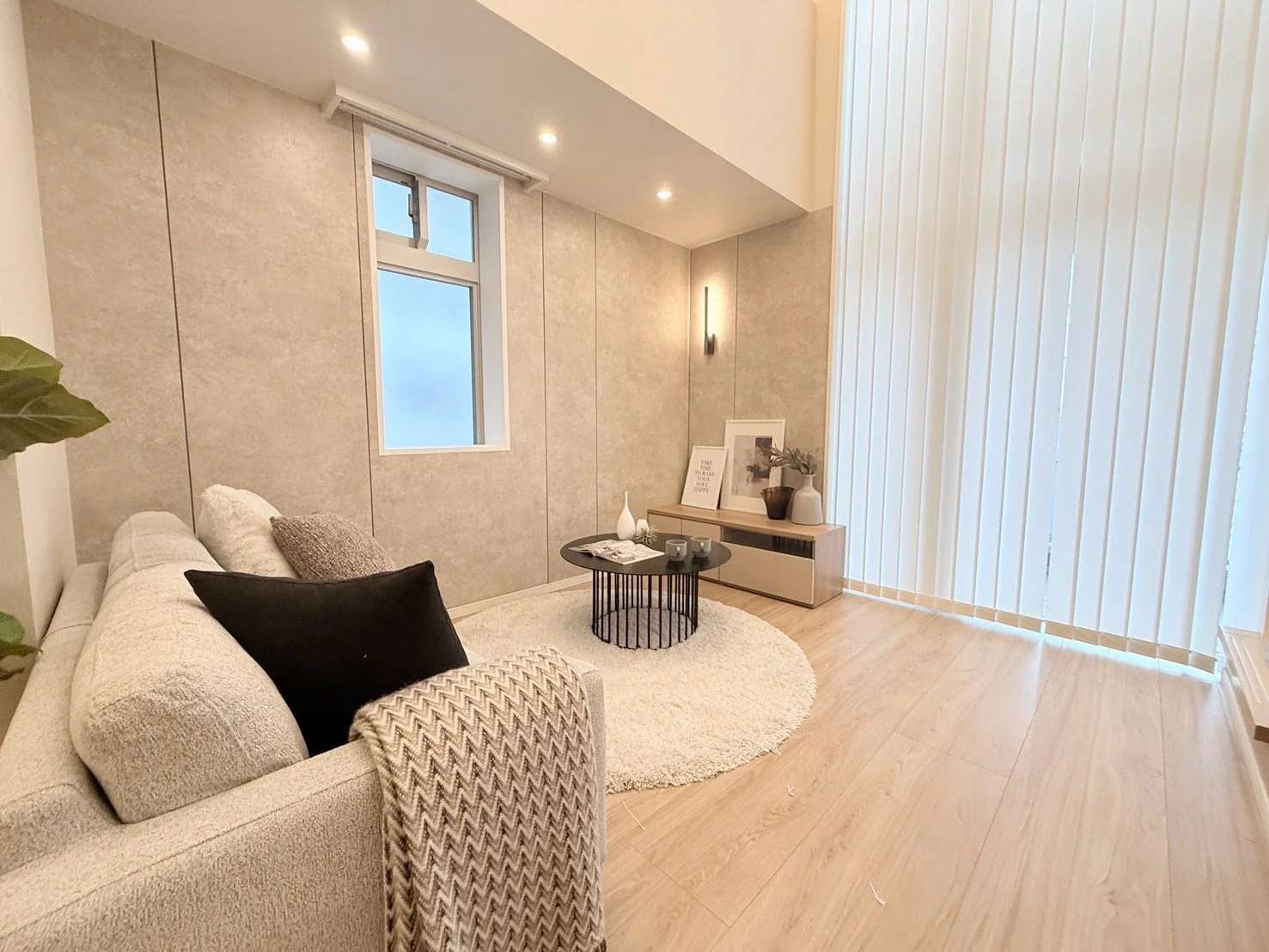 モダンなリビングルームの Interior with beige sofa, black pillow, gray and beige cushion, round white rug, black coffee table, styled with art prints, vases, and potted plants, large window with vertical blinds, wood flooring, recessed ceiling lights, wall-mounte