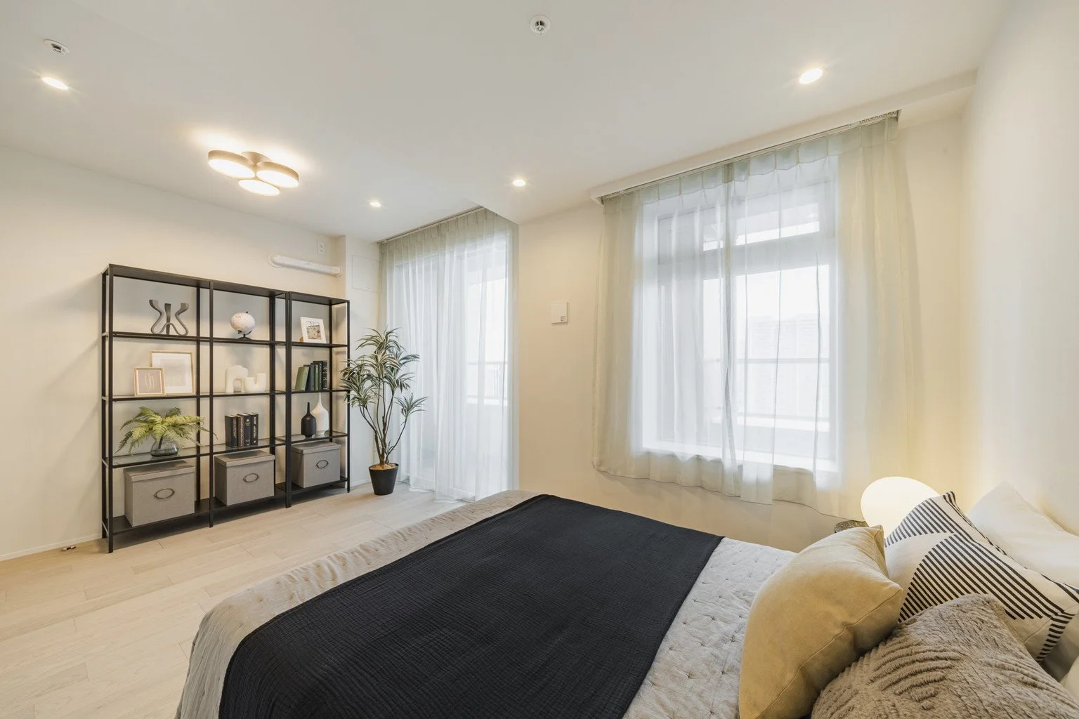 明るい洋室の寝室。ベッドの側に丸いランプ、枕とクッションが置かれている。窓には白いカーテンがかかり、棚には植物や装飾品が飾られている。