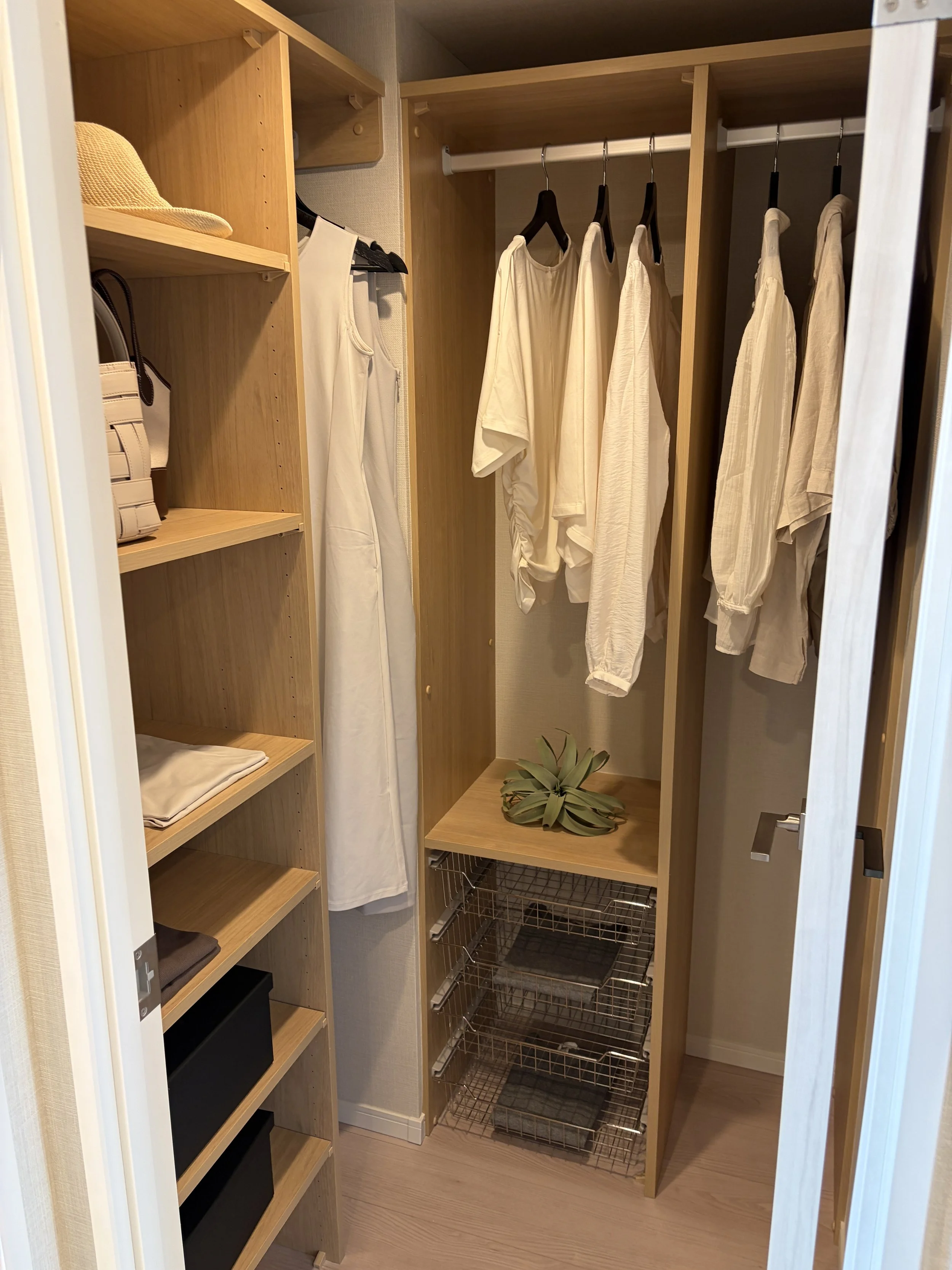 木製のクローゼットに白い衣服とパッションが掛けられている。棚に帽子とバッグ、下に金属の収納ボックスがある。