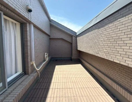 屋上のバルコニーの画像。床にはタイルが敷かれており、側には壁と窓が見える。青空と一部の雲も見える。