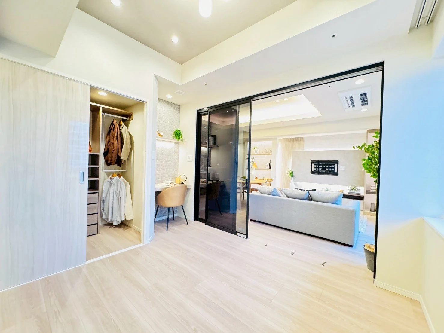 明るいリビングルームとダイニングスペースの写真。仕切りのガラス扉、ソファ、観葉植物、洋服収納棚などが見える。