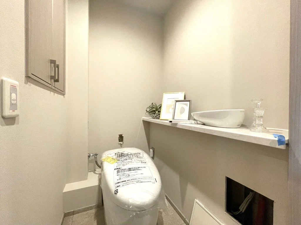 新築のトイレの部屋。便器と棚に飾りや写真、洗面器とガラスのキャンドルスタンドが置かれている。