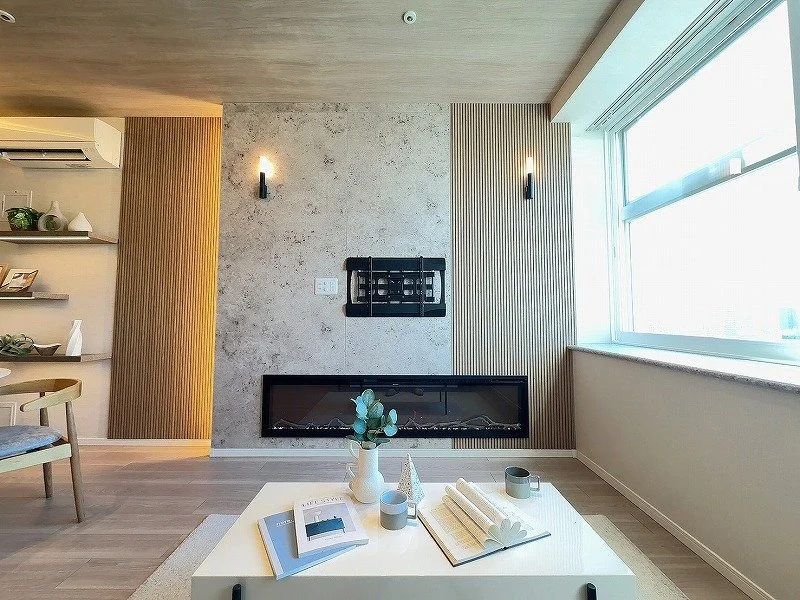 モダンなリビングルーム、中央に白いテーブルと花瓶、右側に大きな窓、壁には暖炉と装飾、左側に棚と椅子。