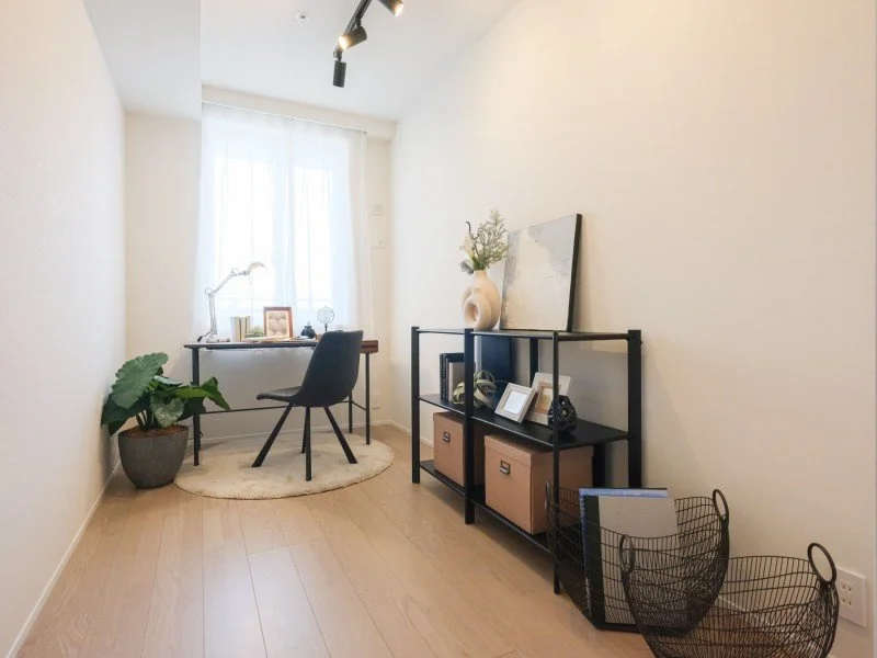 白い壁と木製フローリングの部屋に、小さなデスクと椅子が置かれている。窓から自然光が差し込み、観葉植物や写真立て、本棚が飾られているシンプルなインテリアの部屋。