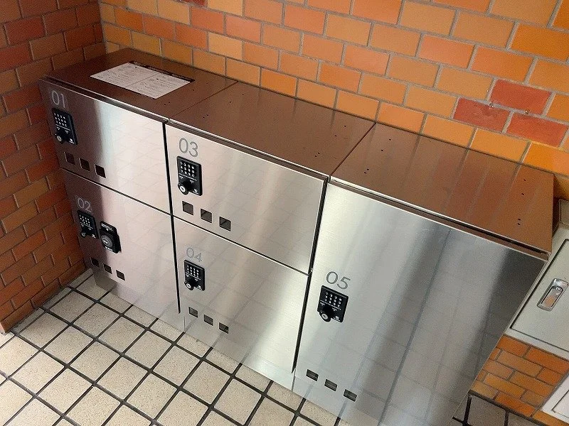 金属製の宅配ボックスが並んでいます。番号は01から05まであり、各ボックスにはエレベーターの暗証番号入力パネルがあります。背景はレンガの壁です。