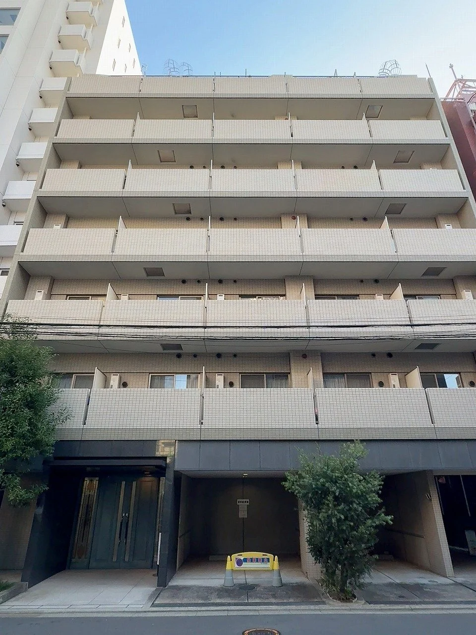 白い多層のマンションの外観。道路から見た建物の正面にはエントランスと駐車場の入り口があり、周囲に木や街路樹がある。