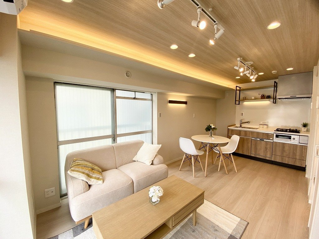 明るく開放的なリビングルームとキッチンの居住空間。白いソファとテーブル。木製の天井と床。自然光と照明が柔らかく照らしている。